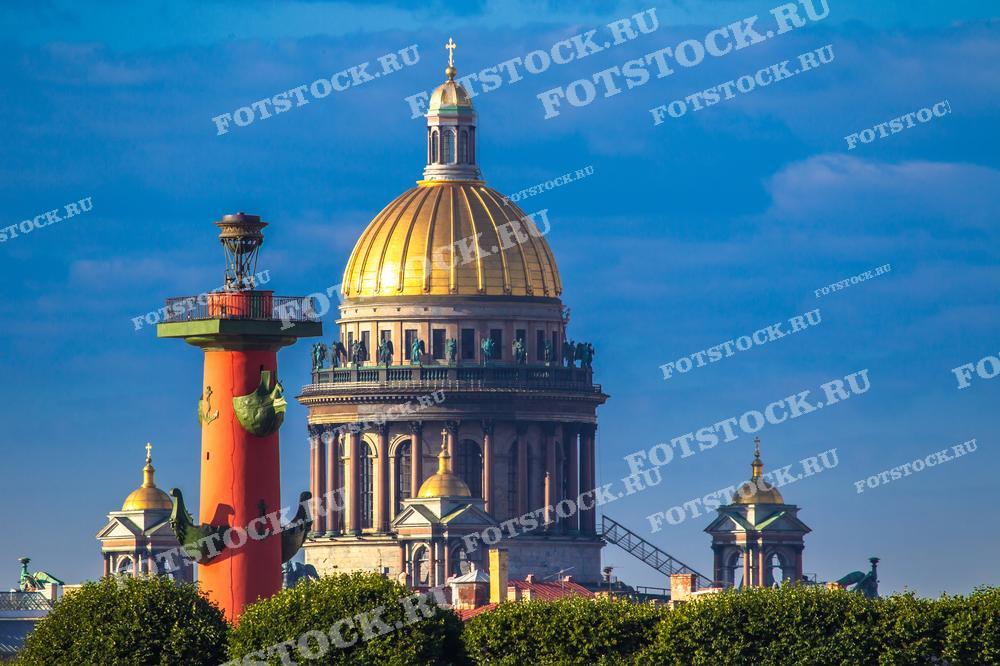 На фотографии изображены Исаакиевский собор и Ростральная колонна в Санкт-Петербурге на фоне синего неба. Виден золотой купол собора и верхняя часть колонны с её украшениями. Фото может использоваться для туристических буклетов, открыток и рекламных материалов.
